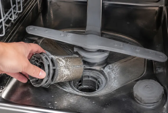 A hand removing a clogged, lime-encrusted filter from the basin of a dishwasher, showing the top signs your home needs immediate appliance repair due to hard water buildup.